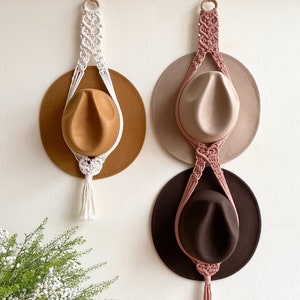 May include: Three hats hanging on a wall using macrame hat holders. The hats are brown, beige, and black. The macrame holders are white, pink, and beige.
