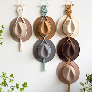 May include: Five hats are hanging on a wall using macrame hat holders. The hats are in various colors, including brown, beige, gray, and white. The macrame holders are made with natural colored cord.