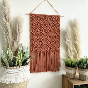 Op de afbeelding: Een roestoranje macramé wandkleed met een geometrisch ruitpatroon en franjes. De wanddecoratie is opgehangen aan een houten stok en een bruin koord. Het wordt getoond met planten in geweven manden en potten.