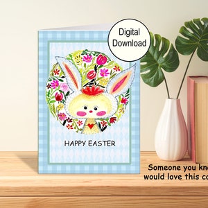 A printable greeting card for Easter. The front of the card has an adorable Easter Bunny I painted for you. The cards says Happy Easter. The card has a blue checked border. This pic shows the card on a table with a vase and books.