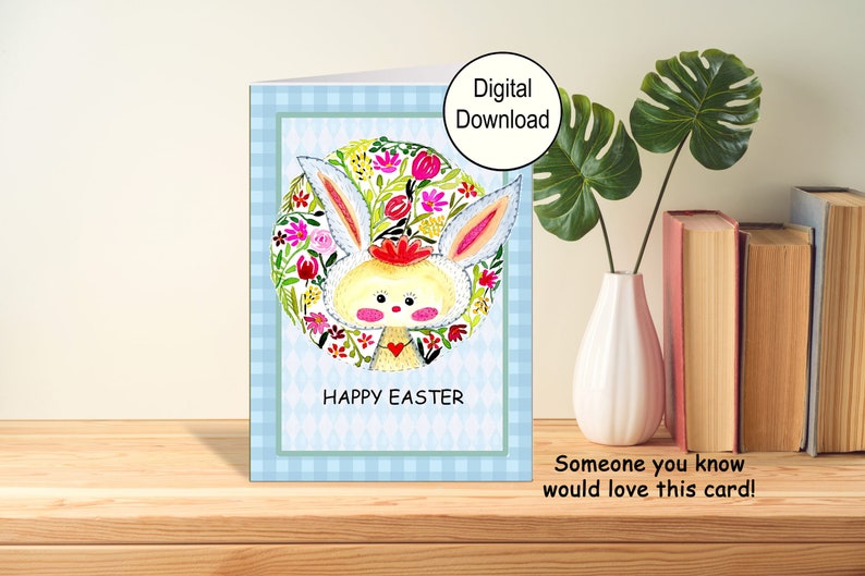A printable greeting card for Easter. The front of the card has an adorable Easter Bunny I painted for you. The cards says Happy Easter. The card has a blue checked border. This pic shows the card on a table with a vase and books.