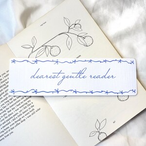May include: A white bookmark with blue decorative trim and the words "dearest gentle reader" in elegant script. The bookmark rests on an open book with botanical illustrations.