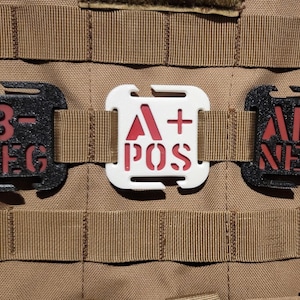 May include: Three blood type identification patches are attached to a tan tactical vest. The patches are black and white with red lettering, displaying blood types B-, A+, and AB-.