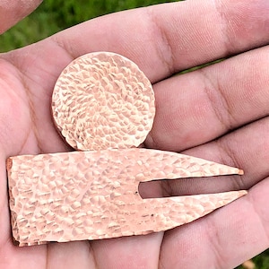 Könnte beinhalten: Ein gehämmerter Golfball-Marker aus Kupfer mit einem passenden, gabelförmigen Divot-Werkzeug.