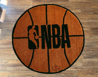 May include: A round brown and black rug with a basketball design. The rug has the NBA logo in the center with a basketball player silhouette. The rug is on a wooden floor.