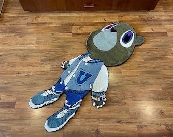 May include: A large, brown and green rug depicting a cartoon bear wearing a blue and white varsity jacket with a large "U" on the front. The bear is lying on its back with its arms outstretched and legs bent at the knees. The rug is on a wooden floor.