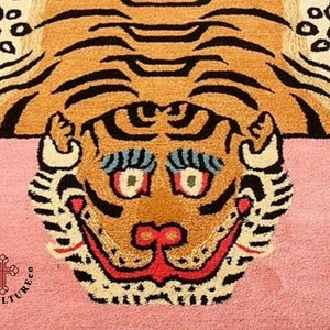K&ouml;nnte beinhalten: Ein rosa Teppich mit einem braunen und schwarzen Tigermuster. Das Gesicht des Tigers ist mittig auf dem Teppich platziert und hat ein breites, zahniges Grinsen. Die Augen des Tigers sind blau und gr&uuml;n, und seine Nase ist rot. Der Teppich hat einen wei&szlig;en Rand um den Rand.