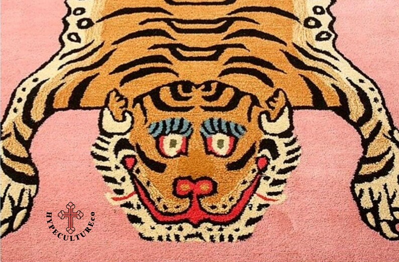 K&ouml;nnte beinhalten: Ein rosa Teppich mit einem braunen und schwarzen Tigermuster. Das Gesicht des Tigers ist mittig auf dem Teppich platziert und hat ein breites, zahniges Grinsen. Die Augen des Tigers sind blau und gr&uuml;n, und seine Nase ist rot. Der Teppich hat einen wei&szlig;en Rand um den Rand.
