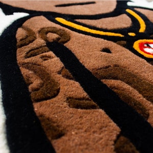 May include: Close-up of a brown and black rug with a detailed design. The rug features a black outline and a brown, textured surface. A yellow and black chain with a red and black circular pendant is visible in the upper right corner.