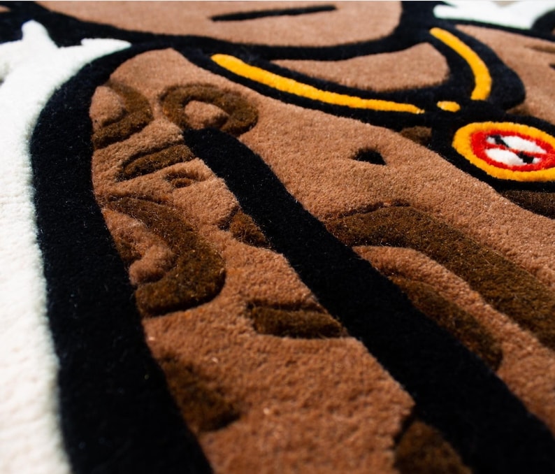 May include: Close-up of a brown and black rug with a detailed design. The rug features a black outline and a brown, textured surface. A yellow and black chain with a red and black circular pendant is visible in the upper right corner.