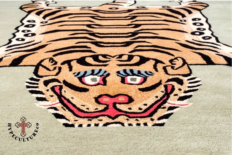 K&ouml;nnte beinhalten: Ein gro&szlig;er Tigerteppich mit gr&uuml;nem Hintergrund. Der Tiger ist braun und schwarz mit einer rosa Nase und einem rosa Maul. Der Teppich hat einen wei&szlig;en Rand und der Text "HYPECULTURE CO" befindet sich in der linken unteren Ecke.