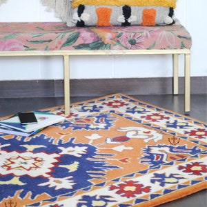 May include: A colorful rug with a geometric pattern in shades of orange, blue, white, and red. The rug is placed on a wooden floor in front of a gold metal bench with a floral patterned cushion.