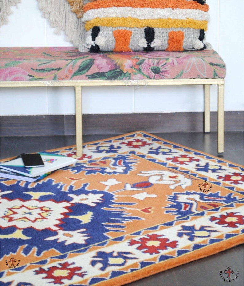 May include: A colorful rug with a geometric pattern in shades of orange, blue, white, and red. The rug is placed on a wooden floor in front of a gold metal bench with a floral patterned cushion.