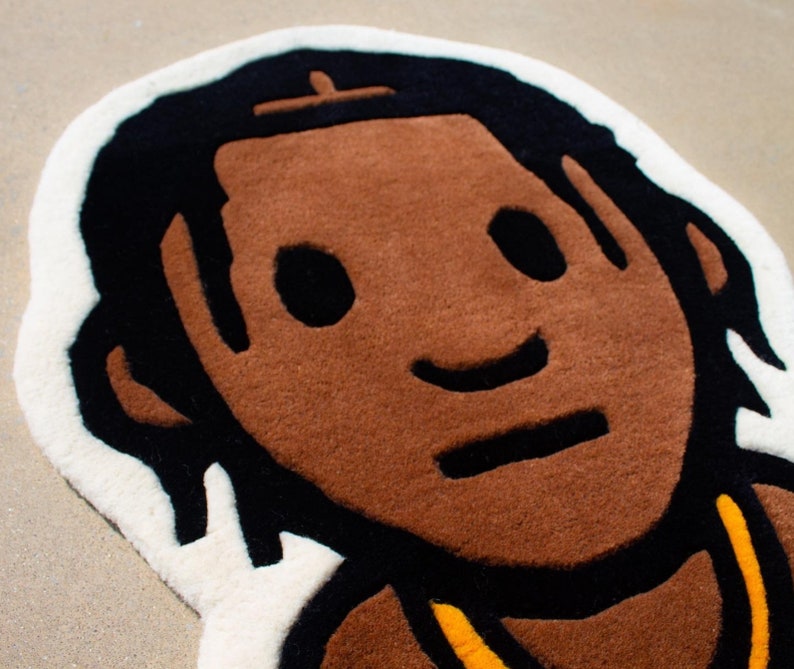 May include: A brown and black rug with a cartoon-style illustration of a person with brown skin, black hair, and a yellow necklace. The person is smiling and has a simple, cartoon-style face.
