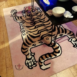 K&ouml;nnte beinhalten: Ein rosa Teppich mit einem Tigermuster in Braun und Schwarz. Der Tiger liegt auf dem R&uuml;cken mit seinen Pfoten in der Luft. Der Teppich hat den Text "HYPECULTURECO" in schwarzen Buchstaben in der linken unteren Ecke.