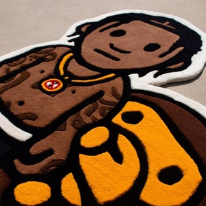 May include: A brown and yellow rug with a cartoon-like image of a person with a gold chain and a black and yellow design. The rug is made of a soft, plush material.