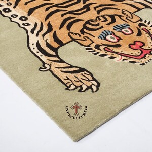 K&ouml;nnte beinhalten: Ein beiger Teppich mit einem braunen und schwarzen Tigerdesign. Der Tiger hat ein breites, zahniges Grinsen und &uuml;bertriebene Augen. Der Teppich ist mit "Hypecultureco" und einem Kreuzsymbol in der rechten unteren Ecke gekennzeichnet.