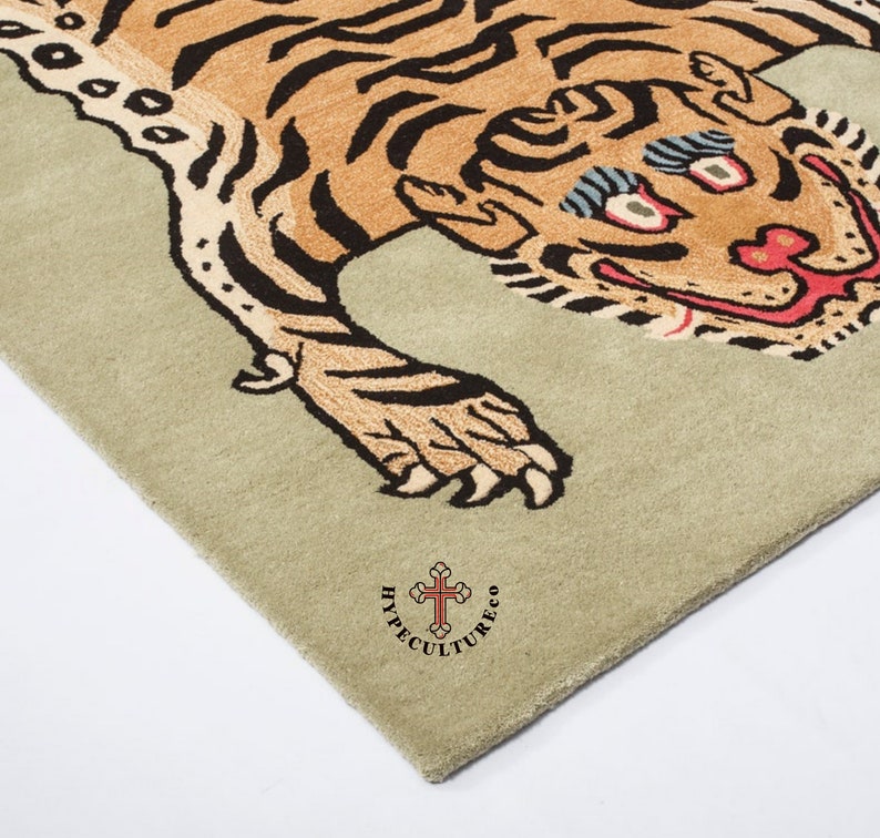 K&ouml;nnte beinhalten: Ein beiger Teppich mit einem braunen und schwarzen Tigerdesign. Der Tiger hat ein breites, zahniges Grinsen und &uuml;bertriebene Augen. Der Teppich ist mit "Hypecultureco" und einem Kreuzsymbol in der rechten unteren Ecke gekennzeichnet.