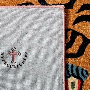K&ouml;nnte beinhalten: Nahaufnahme der R&uuml;ckseite eines Teppichs mit rosa Rand. Der Teppich hat eine graue R&uuml;ckseite mit dem Text "HYPECULTURECO" und einem roten Kreuz in der Mitte.