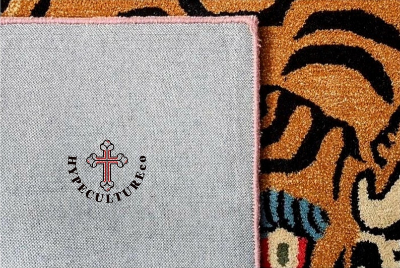 K&ouml;nnte beinhalten: Nahaufnahme der R&uuml;ckseite eines Teppichs mit rosa Rand. Der Teppich hat eine graue R&uuml;ckseite mit dem Text "HYPECULTURECO" und einem roten Kreuz in der Mitte.