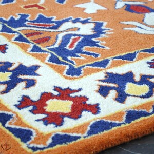 May include: A close-up of a colorful rug with a geometric pattern in shades of orange, blue, white, and red. The rug has a fringe border and is made of wool.