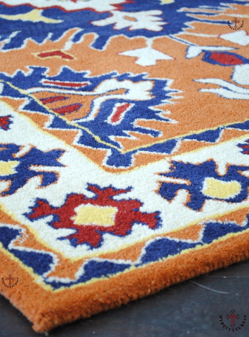 May include: A close-up of a colorful rug with a geometric pattern in shades of orange, blue, white, and red. The rug has a fringe border and is made of wool.