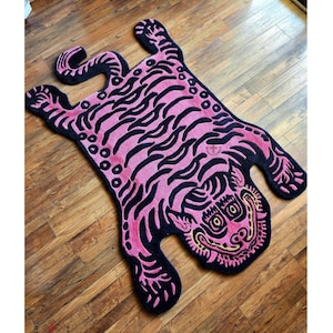 May include: Pink and black tiger rug with a cartoonish design. The rug is made of a soft, plush material and features a detailed tiger face with large eyes.