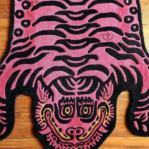 May include: Pink and black rug featuring a tiger face design. The tiger's face is stylised with large eyes and a wide mouth. The rug is made from a soft, plush material.