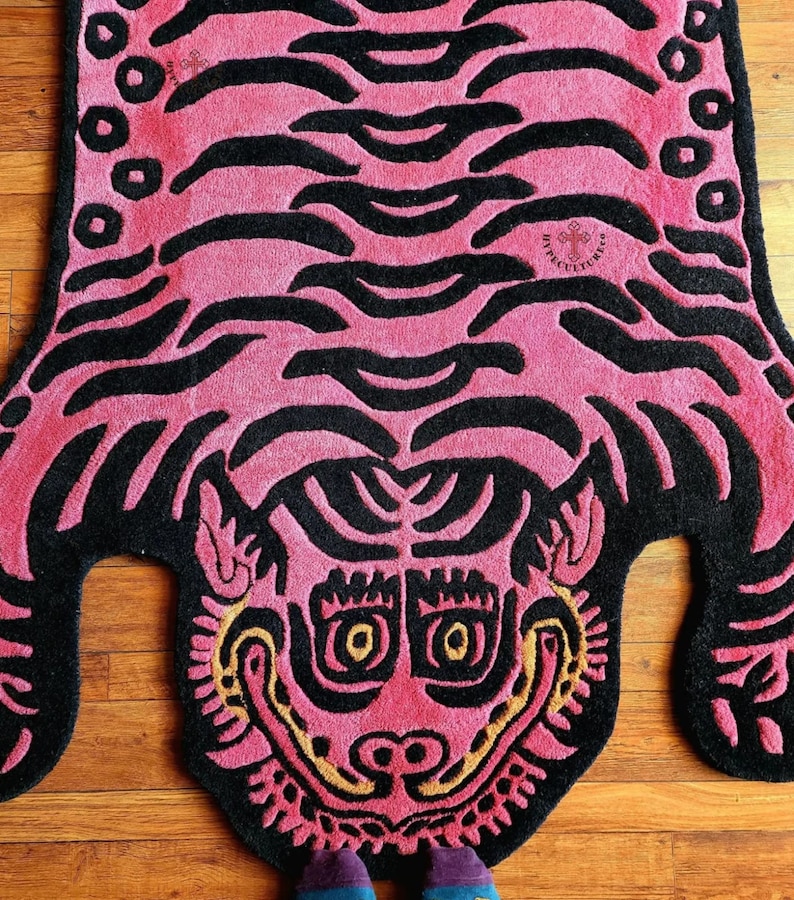 May include: Pink and black rug featuring a tiger face design. The tiger's face is stylised with large eyes and a wide mouth. The rug is made from a soft, plush material.