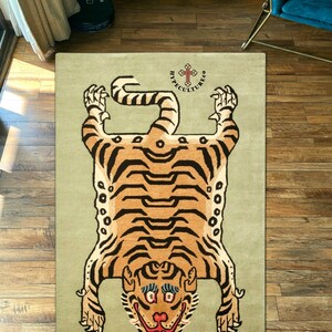 K&ouml;nnte beinhalten: Ein beiger Teppich mit einem Tigerfellmuster. Der Tiger ist braun und schwarz mit einem wei&szlig;en Gesicht. Der Teppich tr&auml;gt den Text "Hypeculture Co." in schwarzen Buchstaben mit einem Kreuz dar&uuml;ber.