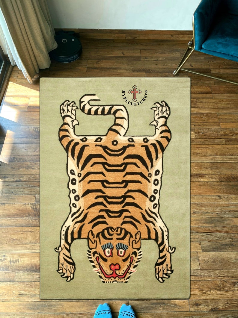 K&ouml;nnte beinhalten: Ein beiger Teppich mit einem Tigerfellmuster. Der Tiger ist braun und schwarz mit einem wei&szlig;en Gesicht. Der Teppich tr&auml;gt den Text "Hypeculture Co." in schwarzen Buchstaben mit einem Kreuz dar&uuml;ber.