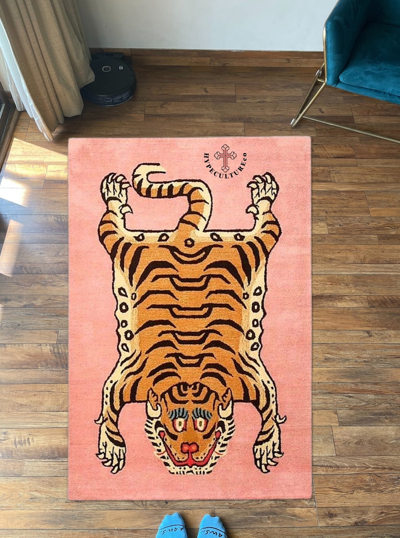 K&ouml;nnte beinhalten: Ein rosa Teppich mit einem Tigerdesign. Der Tiger liegt auf dem R&uuml;cken mit seinen Pfoten in der Luft. Der Teppich tr&auml;gt den Text "HYPECULTURECO" in schwarzen Buchstaben mit einem Kreuzsymbol.