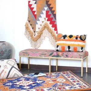 May include: A colorful woven wall tapestry with fringe hangs above a pink floral patterned bench with gold legs. A patterned rug with orange, blue, and white accents is on the floor in front of the bench.