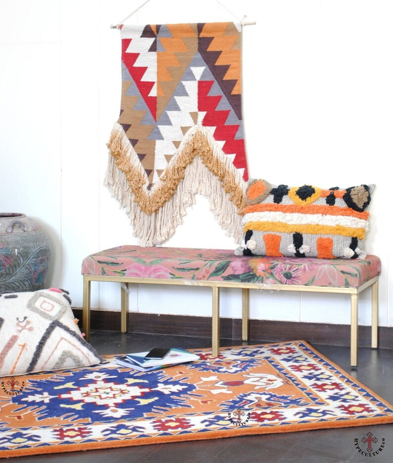 May include: A colorful woven wall tapestry with fringe hangs above a pink floral patterned bench with gold legs. A patterned rug with orange, blue, and white accents is on the floor in front of the bench.