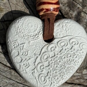 May include: A white heart-shaped pendant with detailed embossed patterns, suspended from a brown suede cord and a striped wooden bead. The pendant is set against a weathered wooden background.