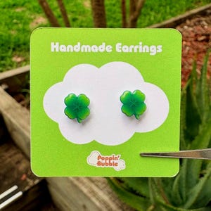 May include: Handmade earrings with bright green four-leaf clover designs. The earrings are presented on a green card with the text "Handmade Earrings" and a white cloud shape. The brand name "Poppin' Bubble" is also visible.