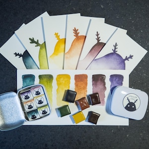 Può includere: Materiali per acquerello tra cui un set di acquerelli in una scatola di metallo, singoli quadrati di vernice e carta per acquerello con disegni di montagne colorate. La scatola ha un logo di cervo dei cartoni animati. La tavolozza dei colori comprende blu, verdi, gialli, marroni e viola.