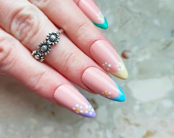Puede incluir: Primer plano de una mano con uñas largas y almendradas pintadas de un color rosa claro. Las puntas de las uñas están pintadas en un degradado de azul, amarillo y verde, con pequeñas flores blancas pintadas en las uñas. Un anillo de plata con tres pequeños diseños de flores está en el dedo anular.