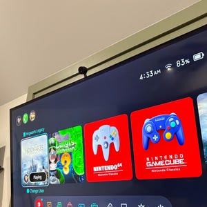 May include: A wide, flat-screen television shows a gaming console interface. The screen displays game icons, including "Hogwarts Legacy," "Luigi's Mansion," and Nintendo console emulators. The time is 4:33 AM, and the battery is at 83%.