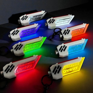 Puede incluir: Una colección de llaveros blancos y negros con un diseño futurista, cada uno emitiendo un color de luz diferente: rojo, amarillo, verde, azul y naranja. El texto "LIVSAC" es visible en la superficie iluminada de cada llavero.