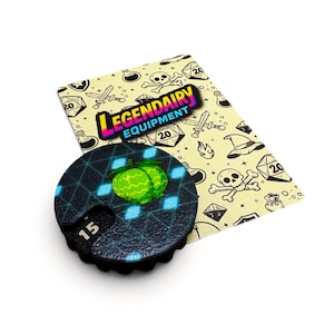 May include: A round black gaming counter with a green fruit graphic and the number 15, resting on a cream-colored card featuring the text LEGENDARY EQUIPMENT and various black line art icons including skulls, swords, and twenty-sided dice.