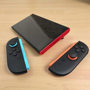 May include: A black and red handheld gaming console with two detached controllers. One controller is black and blue, the other is black and orange. The console is on a light brown wooden surface.