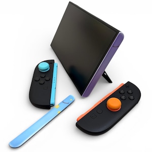 Może przedstawiać: Czarny ekran konsoli do gier z fioletową ramką jest podparty stojakiem. Widoczne są dwa czarne kontrolery z niebieskimi i pomarańczowymi akcentami, a także jasnoniebieski rysik z wzorem gwiazdy. Kontrolery mają przyciski i joysticki.