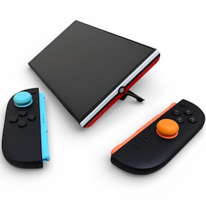 Könnte beinhalten: Eine schwarze Spielkonsole mit roter und weißer Verkleidung, einem schwarzen Bildschirm und einem Ständer. Zwei schwarze Controller mit blauen und orangefarbenen Akzenten und farbigen Tasten sind ebenfalls vorhanden. Die Controller haben einen blauen und orangefarbenen Joystick.