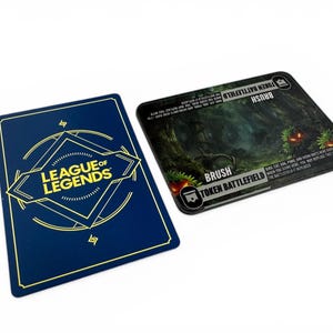 Peut inclure: Deux cartes de jeu League of Legends. L'une est bleu marine avec du texte doré et un logo. L'autre est noire avec une scène de forêt et les mots "BRUSH" et "TOKEN BATTLEFIELD".