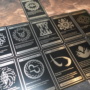 May include: A collection of black and silver cards with various designs and text. The cards feature a variety of symbols and illustrations, including a skull, a flower, and a circle with a line through it. The text on the cards includes the words "Runechant", "Frostbite", "Spectral Shield", "Zen State", and "Ninja Token - Aura".