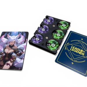 May include: A set of League of Legends game accessories. Includes a card featuring a character illustration, a blue card with the game's logo, and a black tray holding game tokens in purple and green.