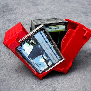 May include: A bright red plastic card holder, open to display a stack of cards. The cards feature images and text, including "SECURITY COMPLEX" and "+KANAN JARRUS". The holder is on a gray surface.