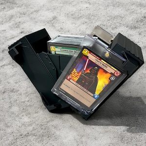 Könnte beinhalten: Schwarze, offene Kartenaufbewahrungsbox mit Sammelkarten. Die Box enthält einen Stapel Karten und eine Karte mit Darth Vader. Die Karten befinden sich in Schutzhüllen. Die Box steht auf einer grauen Oberfläche.