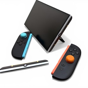 May include: A black gaming console with a black screen and silver trim is propped up with a stand. Two black controllers with colored accents and a black and white accessory are also displayed. The controllers have blue and orange accents.